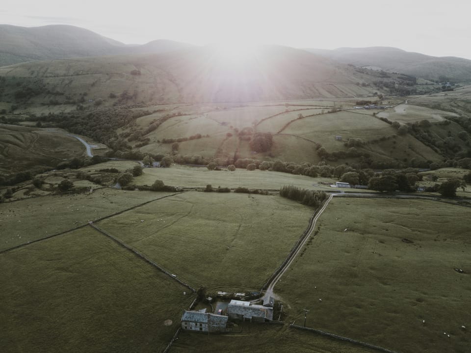 Aerial view of green hills and farmland at dawn. Stone farm buildings sit near winding roads and dry-stone walls, as soft light breaks over the distant horizon.